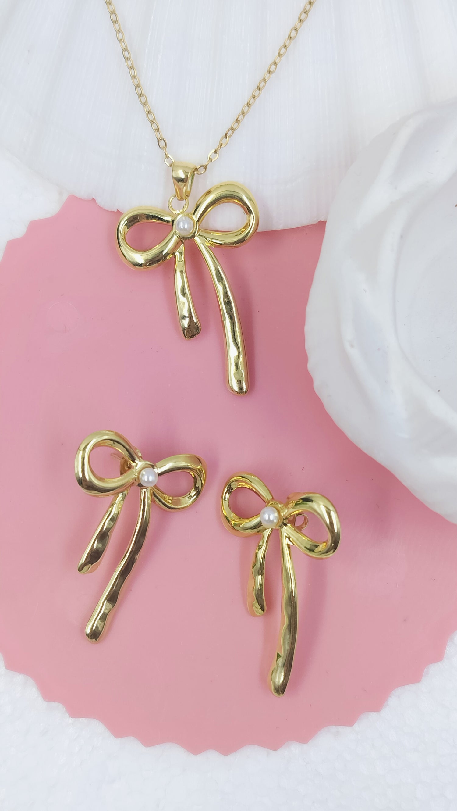 A set of gold-colored bow knot pendant jewelry, including a necklace and earrings, displayed on a pink surface.