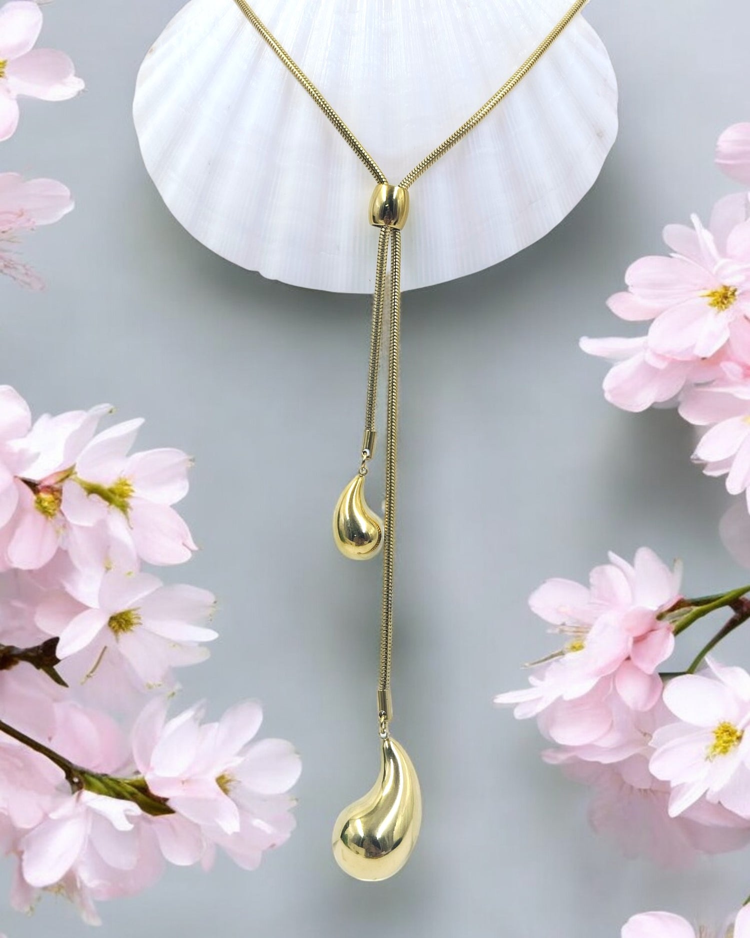 A gold-colored long Y chain necklace with a droplet pendant, displayed against a background with pink floral elements.