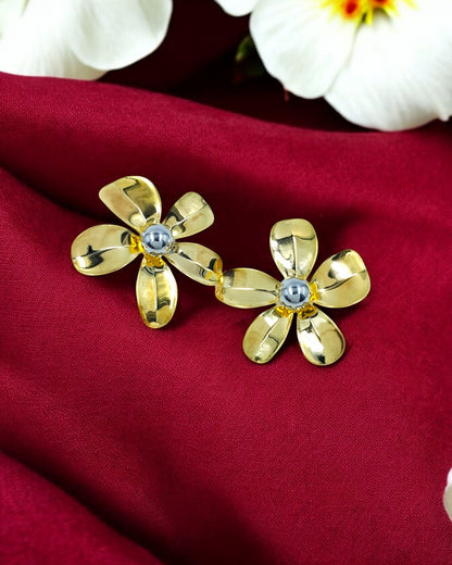 A pair of gold-tone large flower stud statement earrings with a diamond-like embellishment at the center, displayed on a red fabric background with white flowers.
