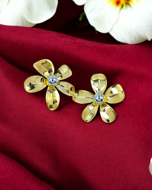 A pair of gold-tone large flower stud statement earrings with a diamond-like embellishment at the center, displayed on a red fabric background with white flowers.
