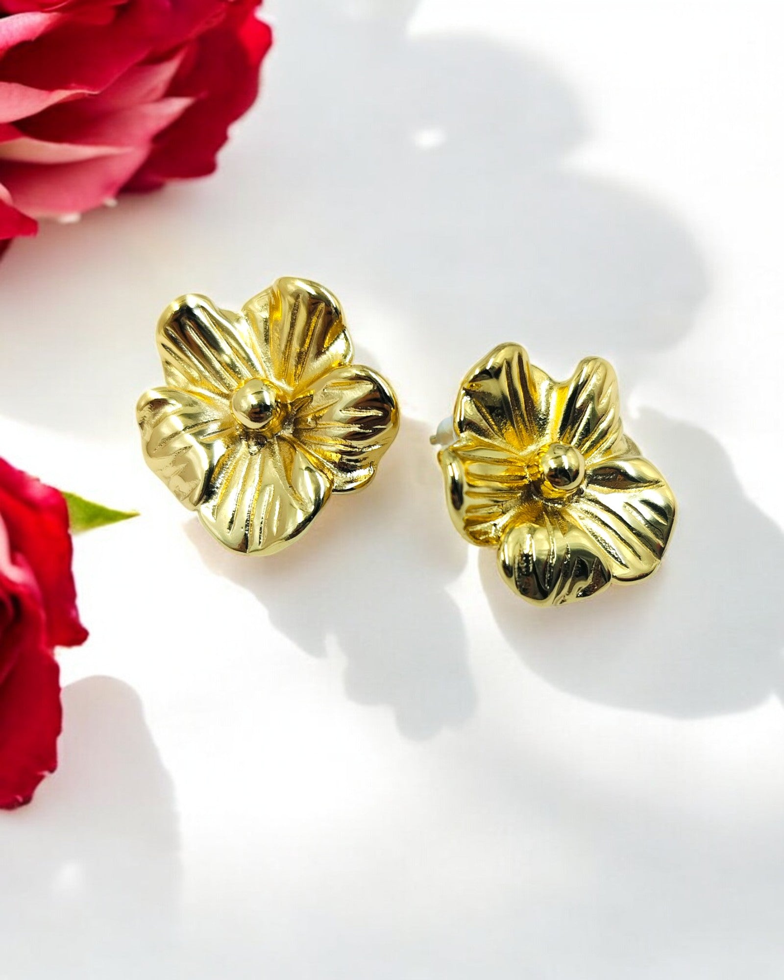 A pair of gold-colored floral stud earrings displayed on a white surface with red flowers in the background.