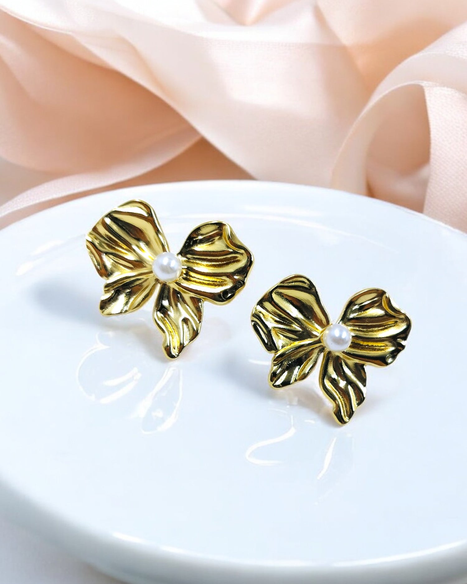A pair of gold-colored bow earrings with pearl accents, displayed on a white plate with a satin fabric background.