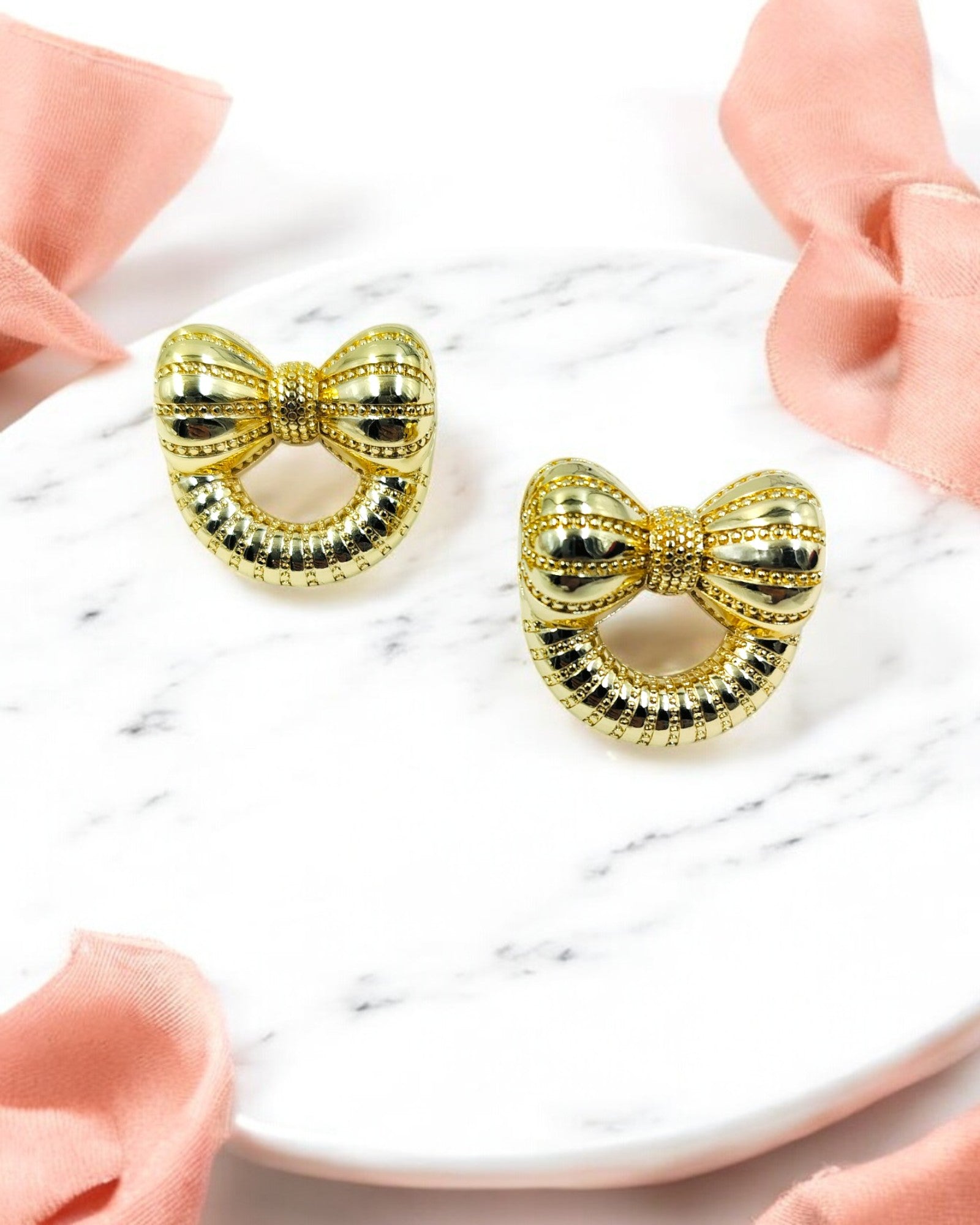 A pair of gold-colored bow-shaped earrings on a white surface with a pink ribbon beside them.