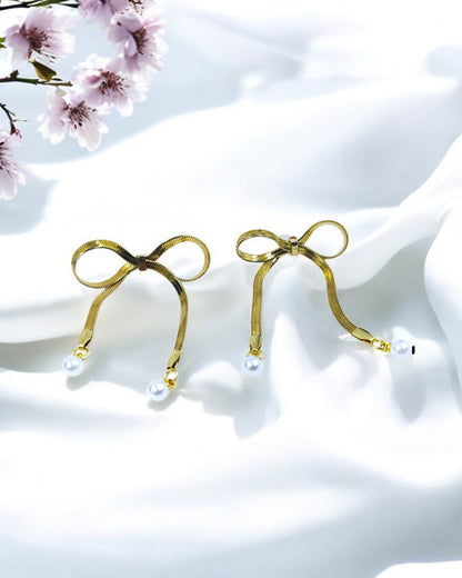 A pair of gold-plated bow chain tassel earrings with a pearl center, displayed against a satin background with floral accents.