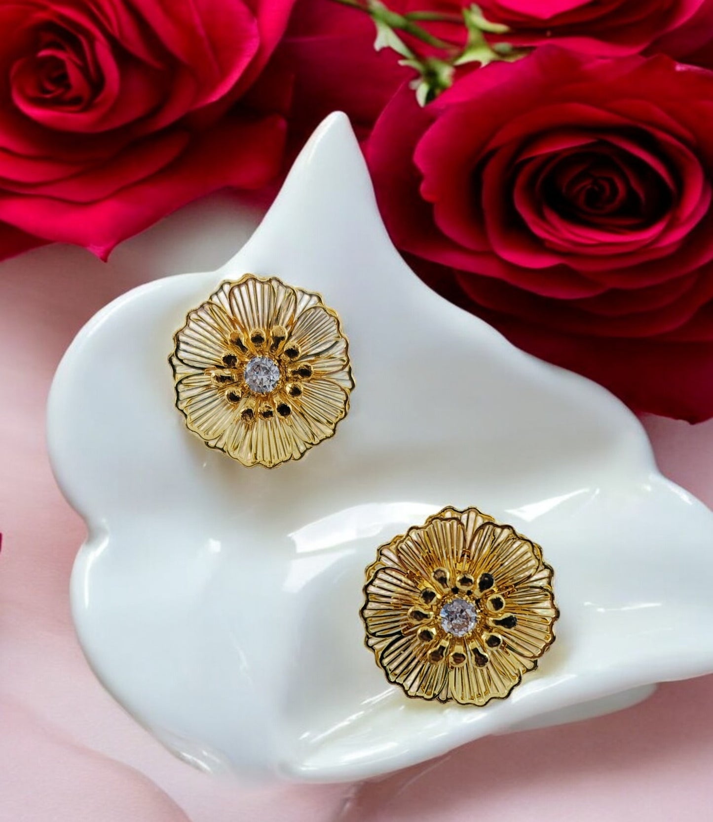 A pair of gold-colored stud earrings with a hibiscus flower design, displayed on a white dish, with red roses in the background.