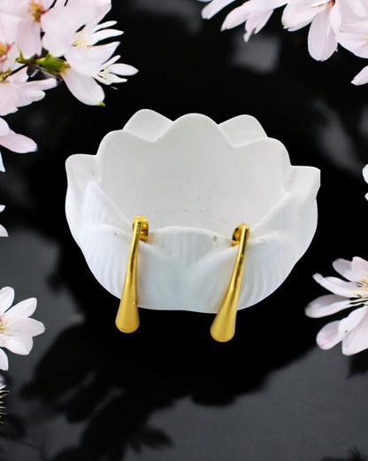 Modern gold-plated earrings with a curved, polished drop shape, sitting in a white ceramic bowl on a black surface with flowers.