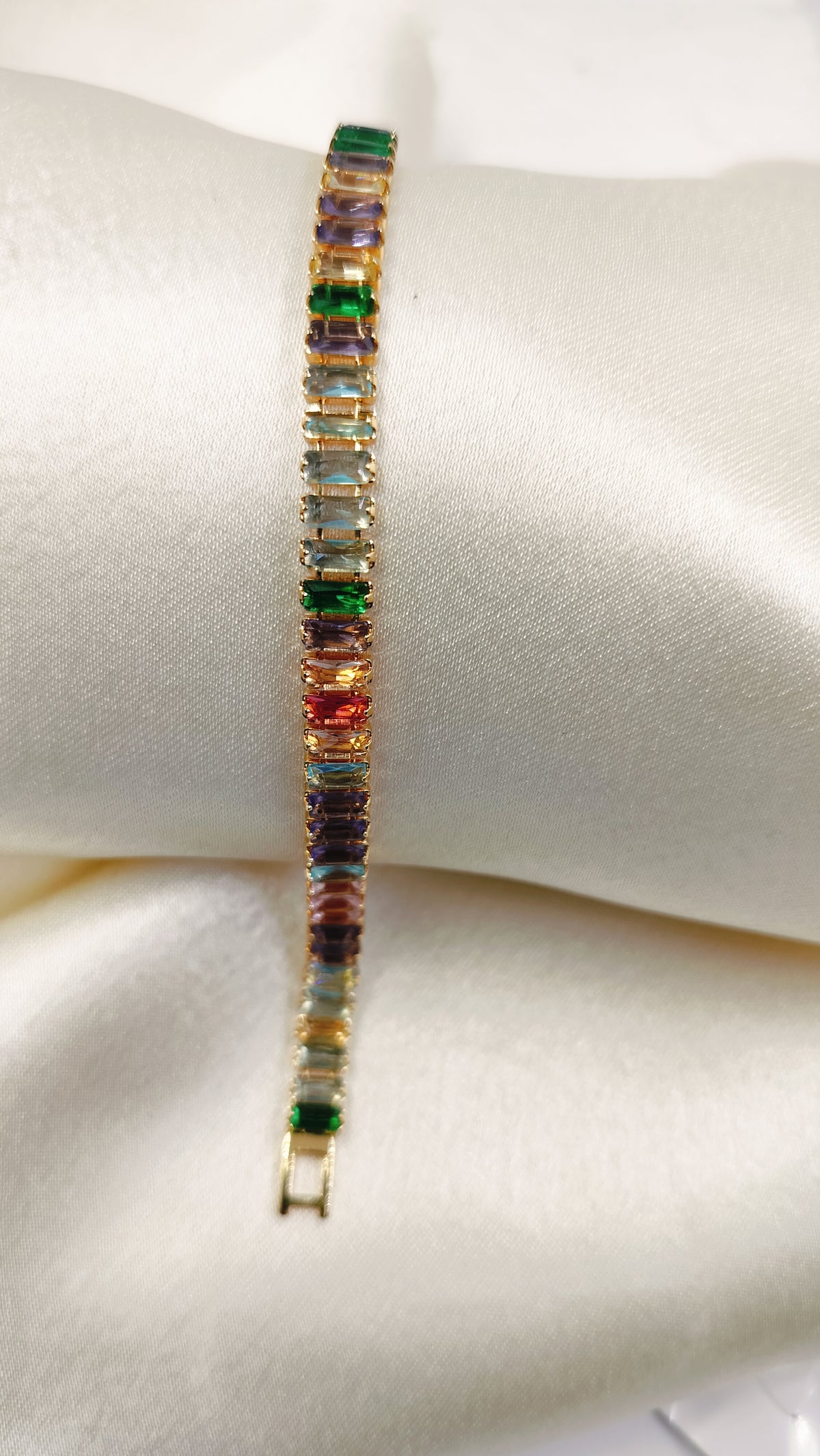 Gold tennis bracelet with multicolored baguette-cut cubic zirconia stones on a satin pillow.