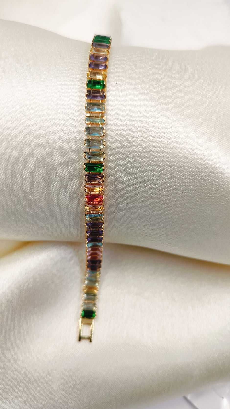 Gold tennis bracelet with multicolored baguette-cut cubic zirconia stones on a satin pillow.