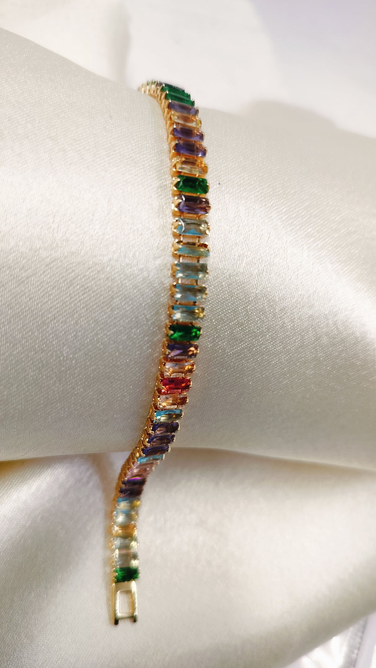 Gold tennis bracelet with multicolored baguette-cut cubic zirconia stones on a satin background.