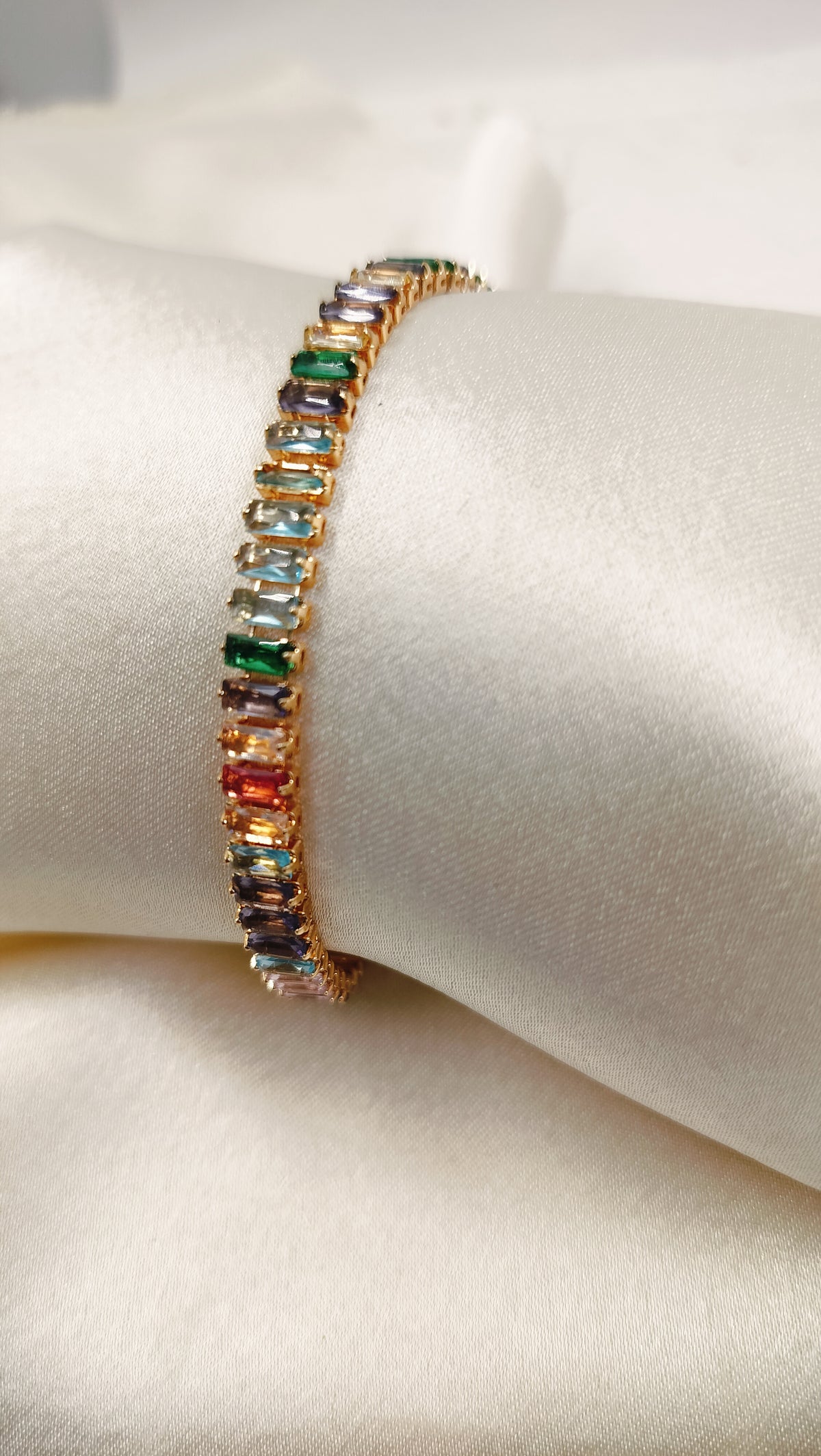 A gold-plated tennis bracelet featuring multi-colored cubic zirconia stones displayed on a white surface.