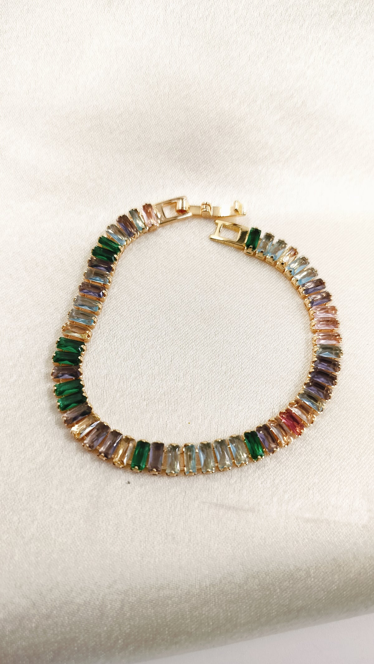Elegant gold-toned bracelet featuring a vibrant row of multicolored rectangular gemstones, displayed on a textured white background.