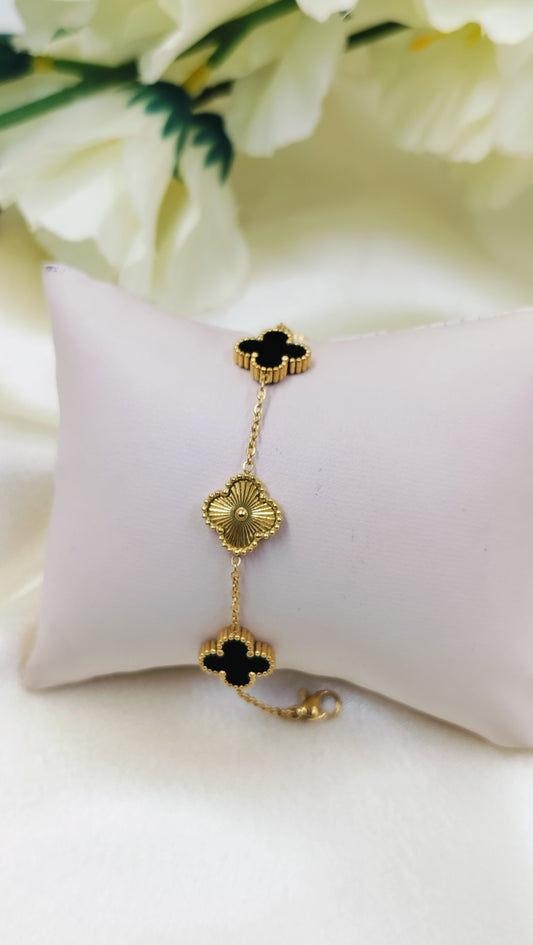A gold-plated bracelet with black textured clover charms, displayed on a white cushion.