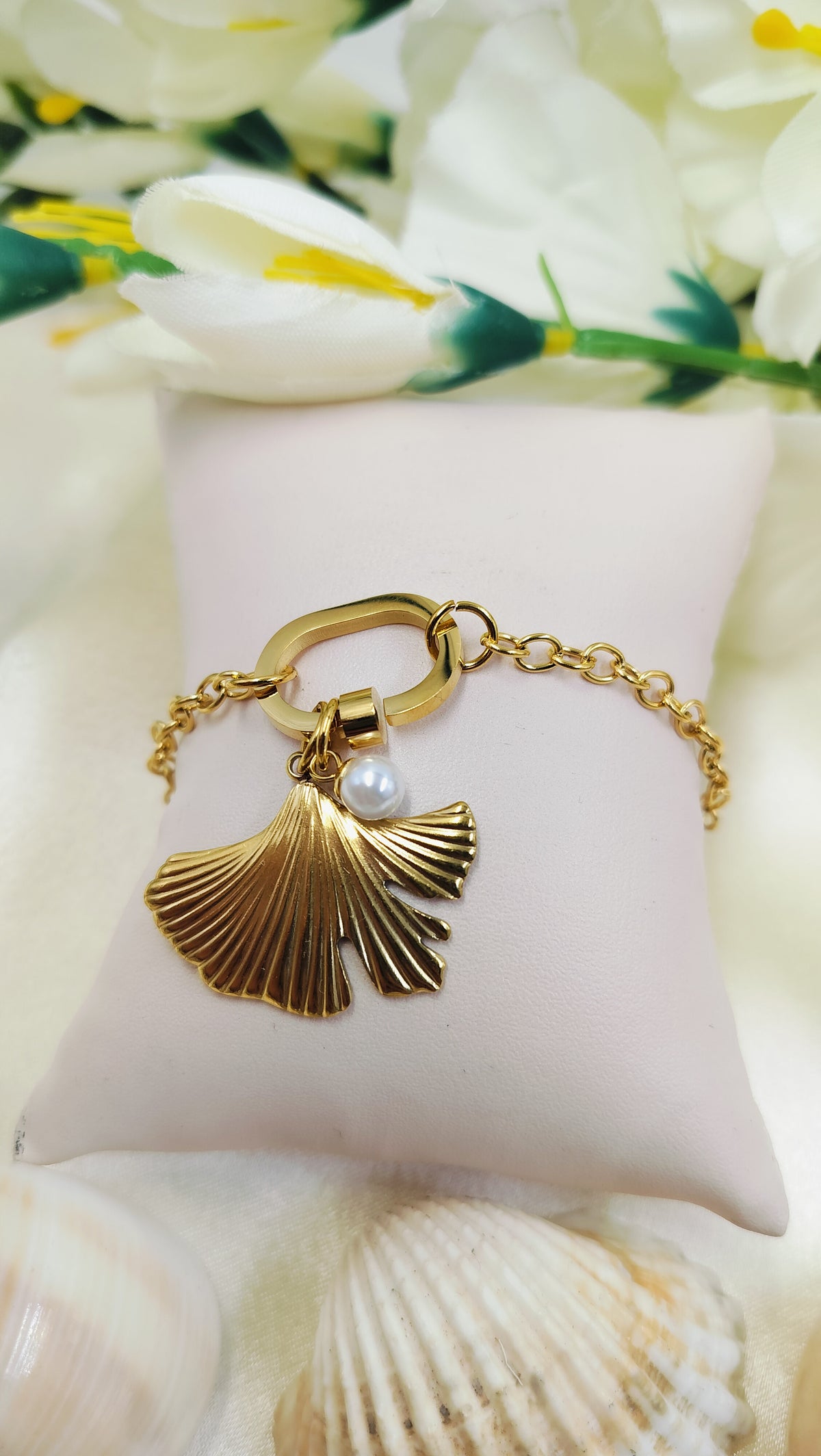 A gold-plated bracelet with a palm leaf charm and a pearl, displayed on a white cushion with floral patterns in the background.