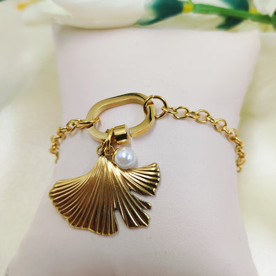 A gold-plated bracelet with a palm leaf charm and a pearl, displayed on a white cushion with floral patterns in the background.