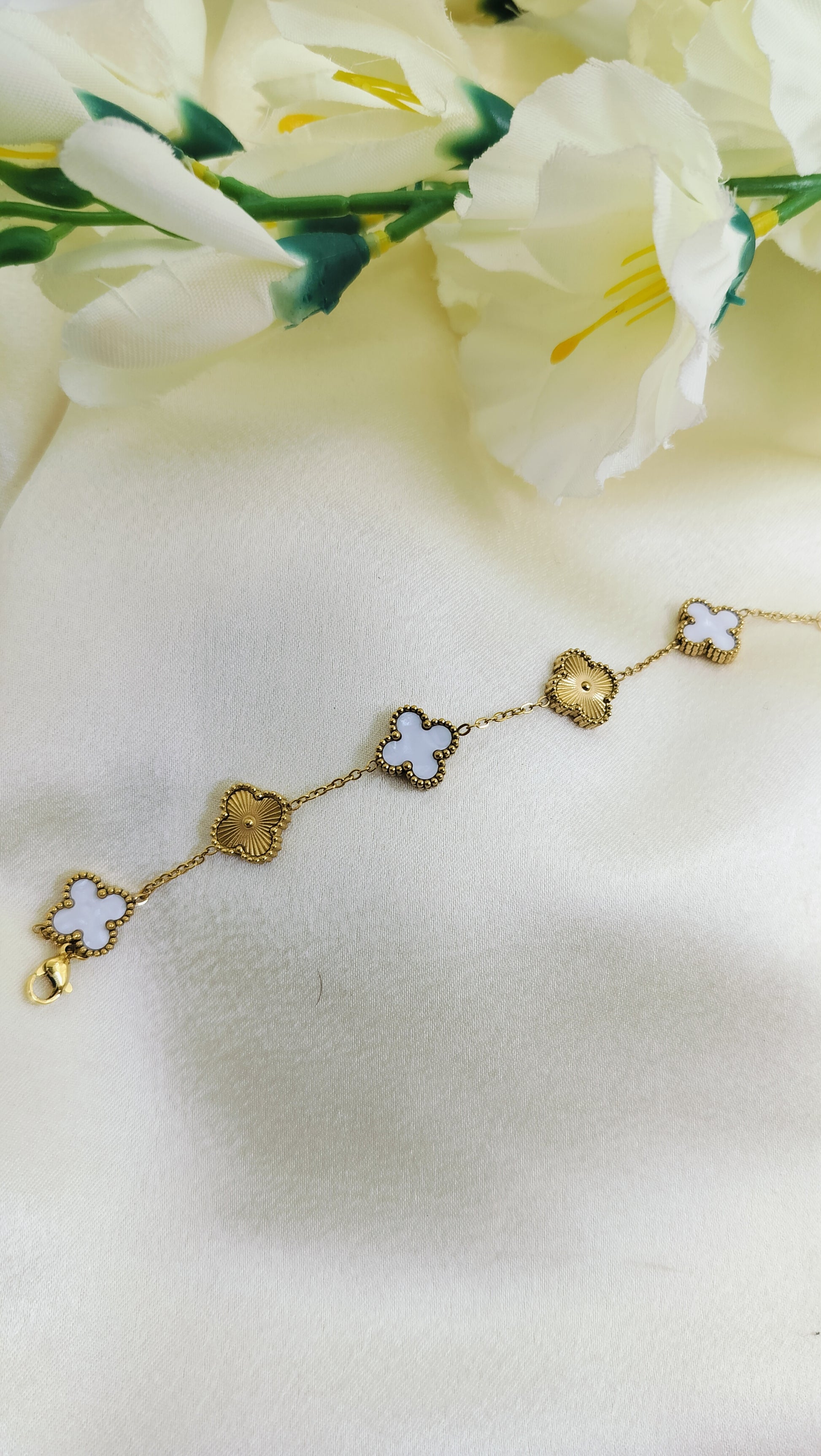 A gold-plated bracelet with white textured clover charms displayed on a silky white surface with flowers in the background.