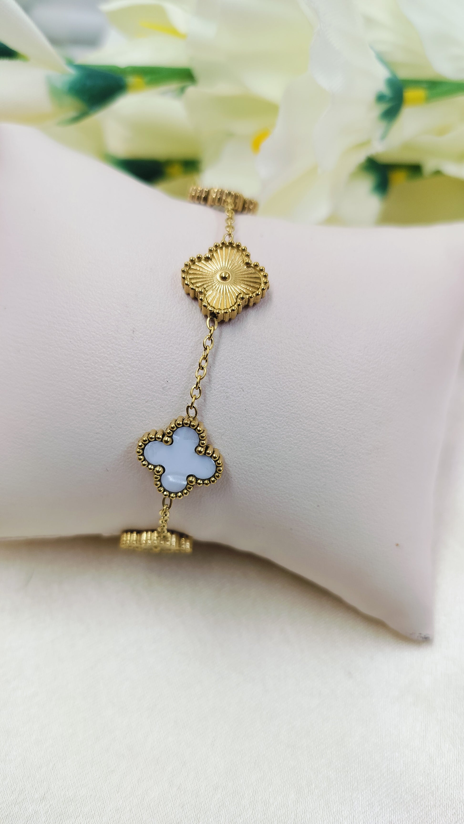 Gold chain bracelet with floral motifs and blue clover charm, displayed on cushion – elegant designer jewelry for women