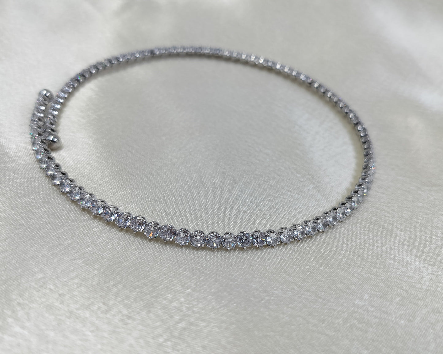 A close-up image of a silver choker necklace encrusted with cubic zirconia crystals, displayed on a white satin surface.