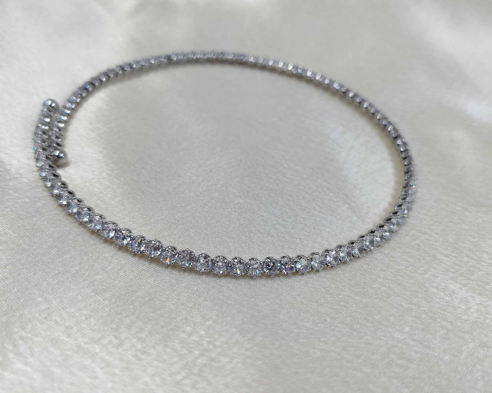 A close-up image of a silver choker necklace encrusted with cubic zirconia crystals, displayed on a white satin surface.