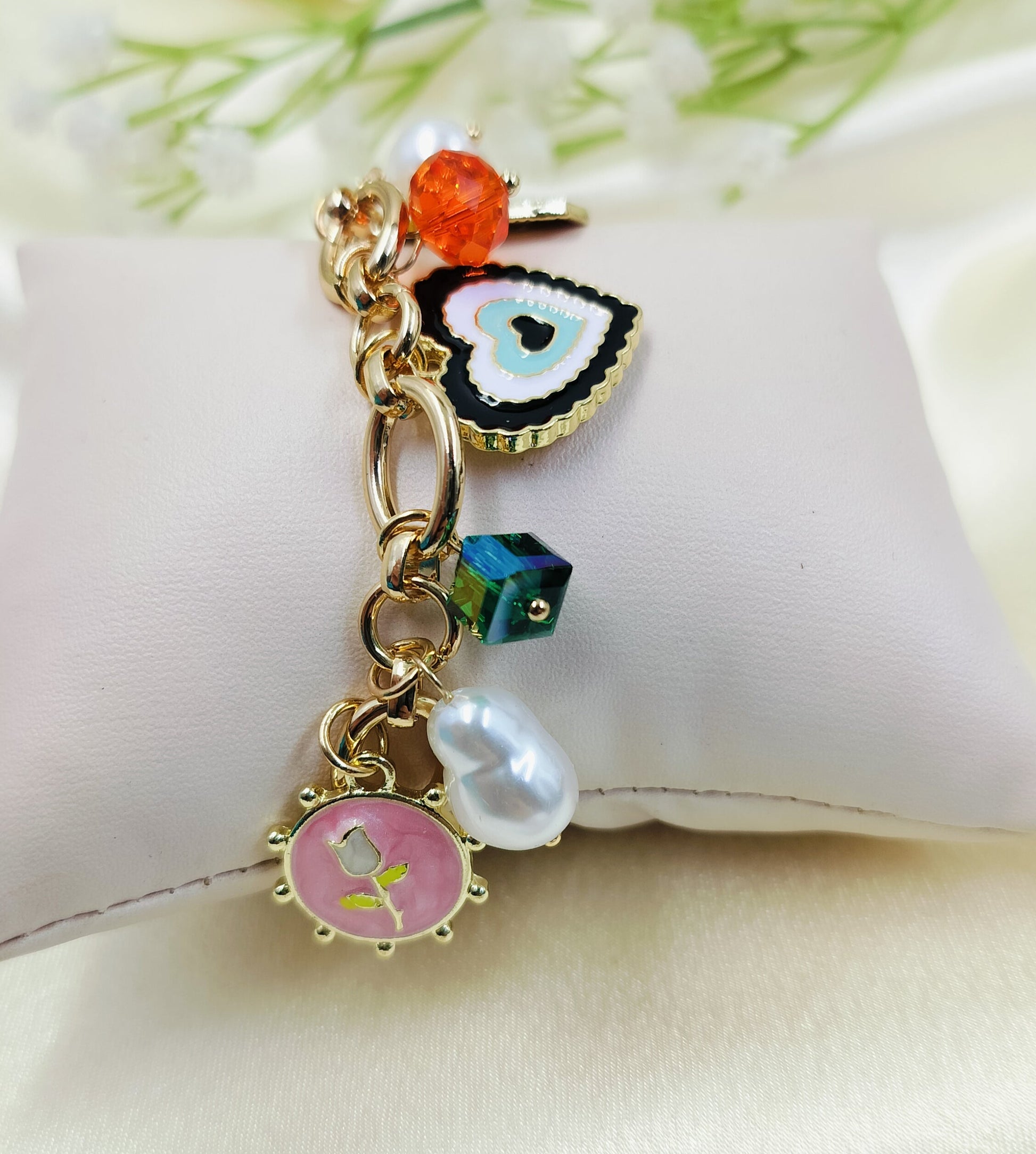 Gold chain bracelet with colorful charms including pink floral enamel, pearl drop, green cube bead, black heart pendant, and orange crystal accent, stylish women’s jewelry on satin fabric cushion background