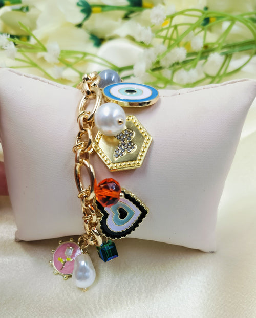 Gold chain bracelet with assorted charms including hexagon pendant, black heart charm, evil eye design, orange crystal bead, pearl accents, and pink floral enamel charm, fashionable women’s jewelry on satin fabric cushion background