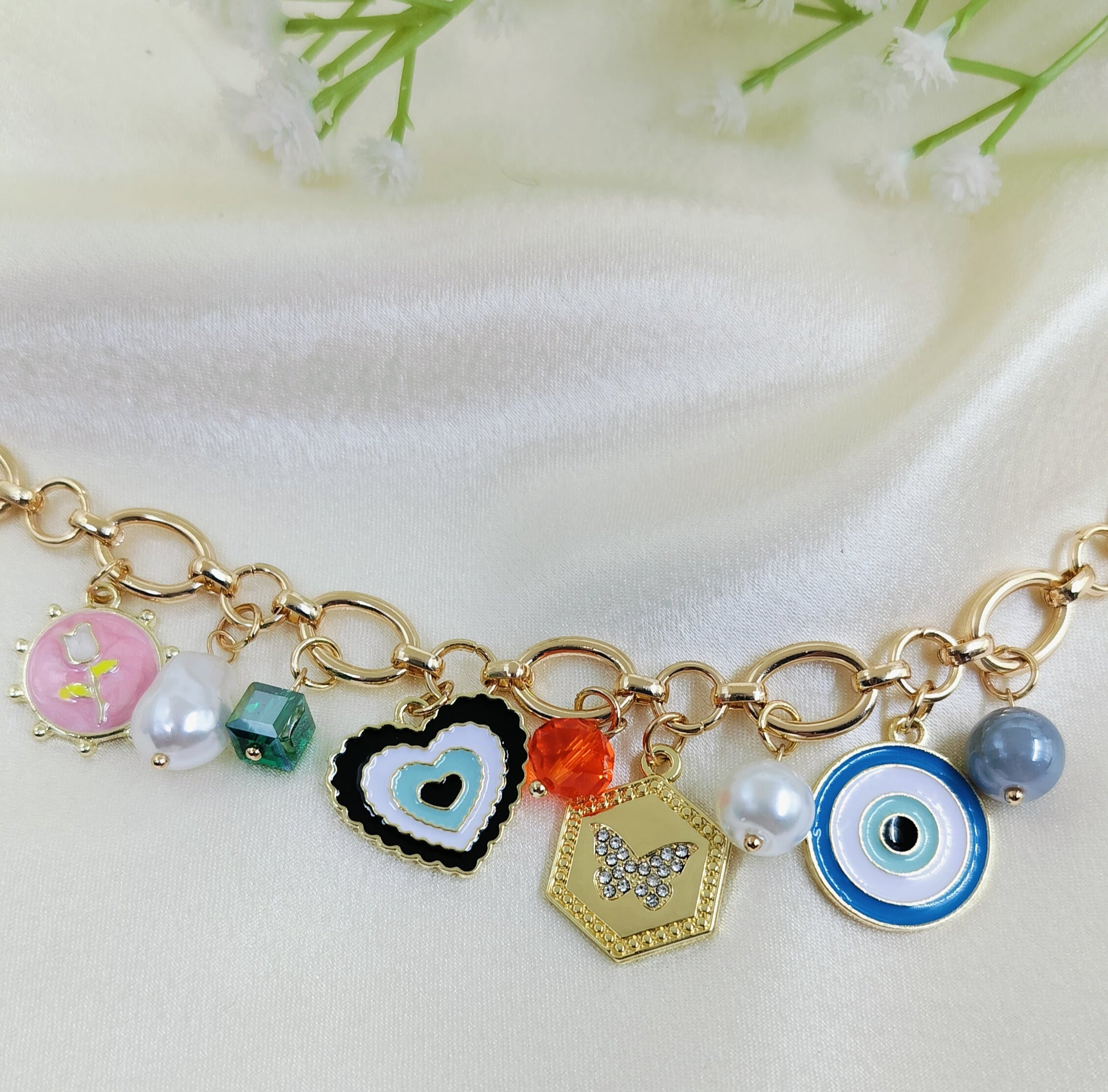 Gold chain bracelet with colorful charms including pink floral enamel, green bead, black heart pendant, orange crystal bead, hexagon butterfly pendant, pearl accents, and blue evil eye design, trendy women’s jewelry on satin fabric background
