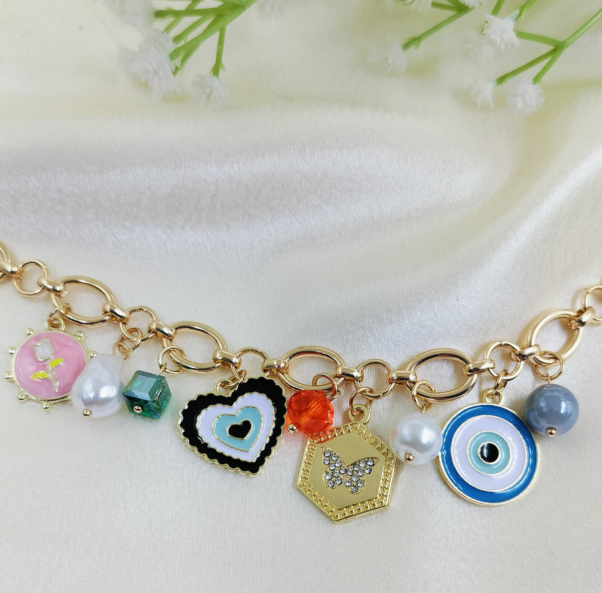 Gold chain bracelet with colorful charms including pink floral enamel, green bead, black heart pendant, orange crystal bead, hexagon butterfly pendant, pearl accents, and blue evil eye design, trendy women’s jewelry on satin fabric background