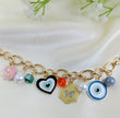 Gold chain bracelet with colorful charms including pink floral enamel, green bead, black heart pendant, orange crystal bead, hexagon butterfly pendant, pearl accents, and blue evil eye design, trendy women’s jewelry on satin fabric background