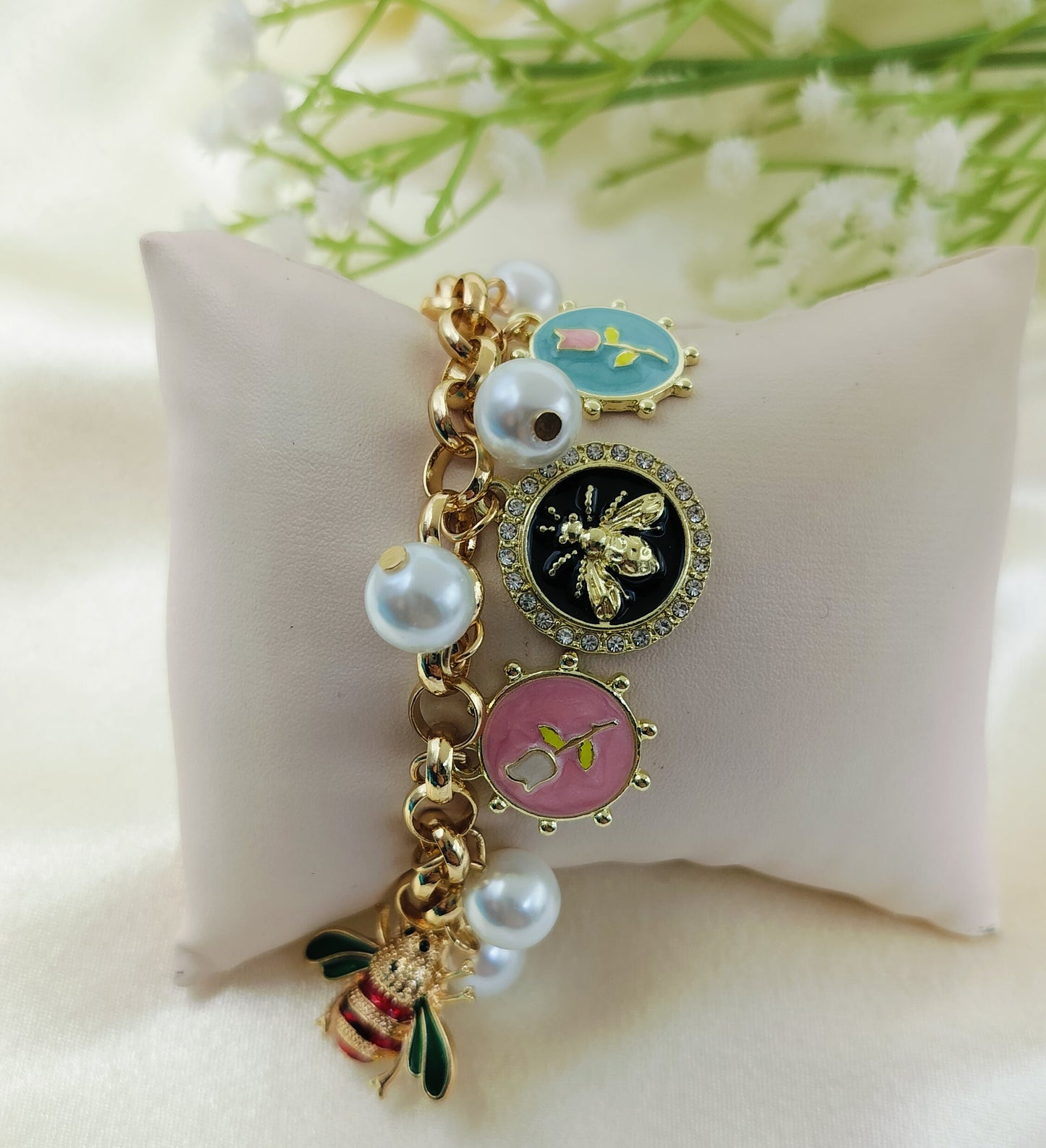 Gold charm bracelet with enamel bee, floral motifs in blue, black, and pink, accented with pearl charms, trendy women’s jewelry on satin fabric cushion background