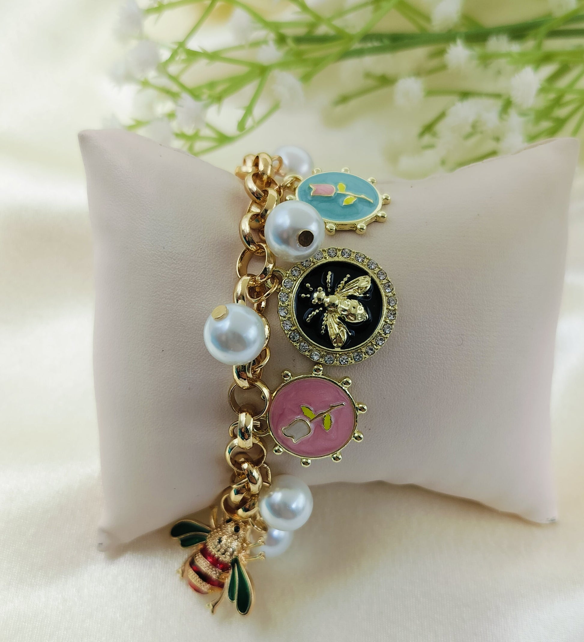 Gold charm bracelet with enamel bee, floral motifs in blue, black, and pink, accented with pearl charms, trendy women’s jewelry on satin fabric cushion background