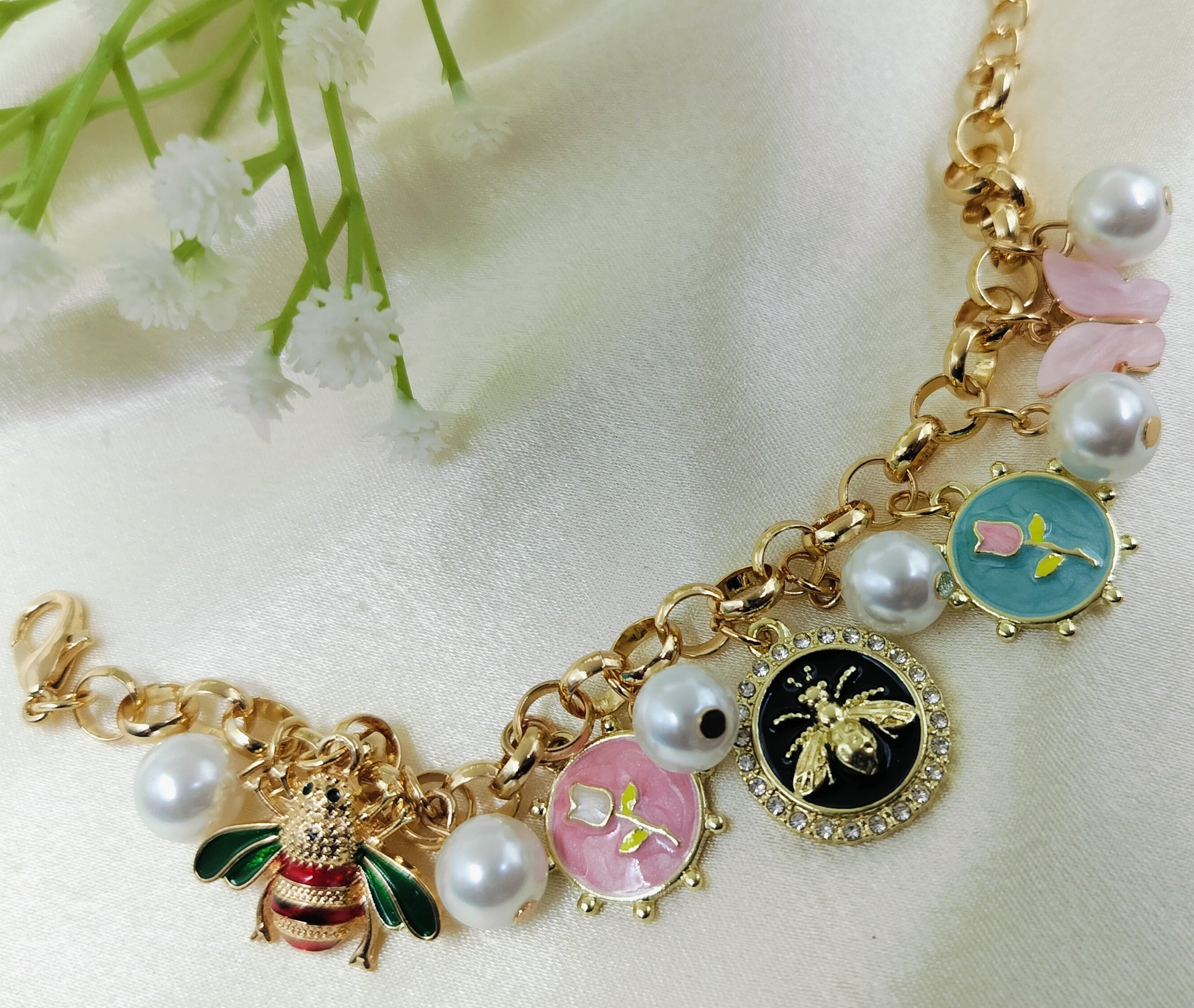 Gold chain bracelet with enamel bee charm, floral motifs in pink, blue, and black, accented with pearls and crystal details, fashionable women’s jewelry on satin fabric background