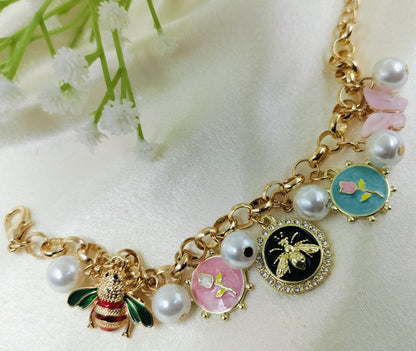 Gold chain bracelet with enamel bee charm, floral motifs in pink, blue, and black, accented with pearls and crystal details, fashionable women’s jewelry on satin fabric background