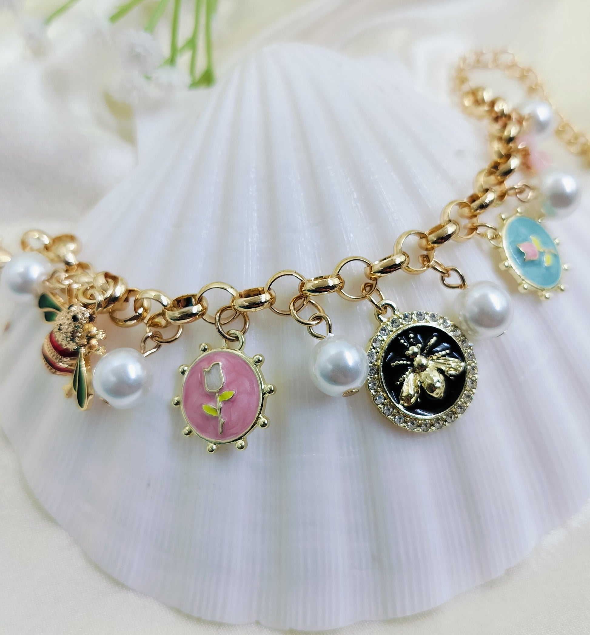 A gold charm bracelet with floral and animal-inspired charms, including a bee, a butterfly, and various pastel-colored medallions, displayed on a white surface.