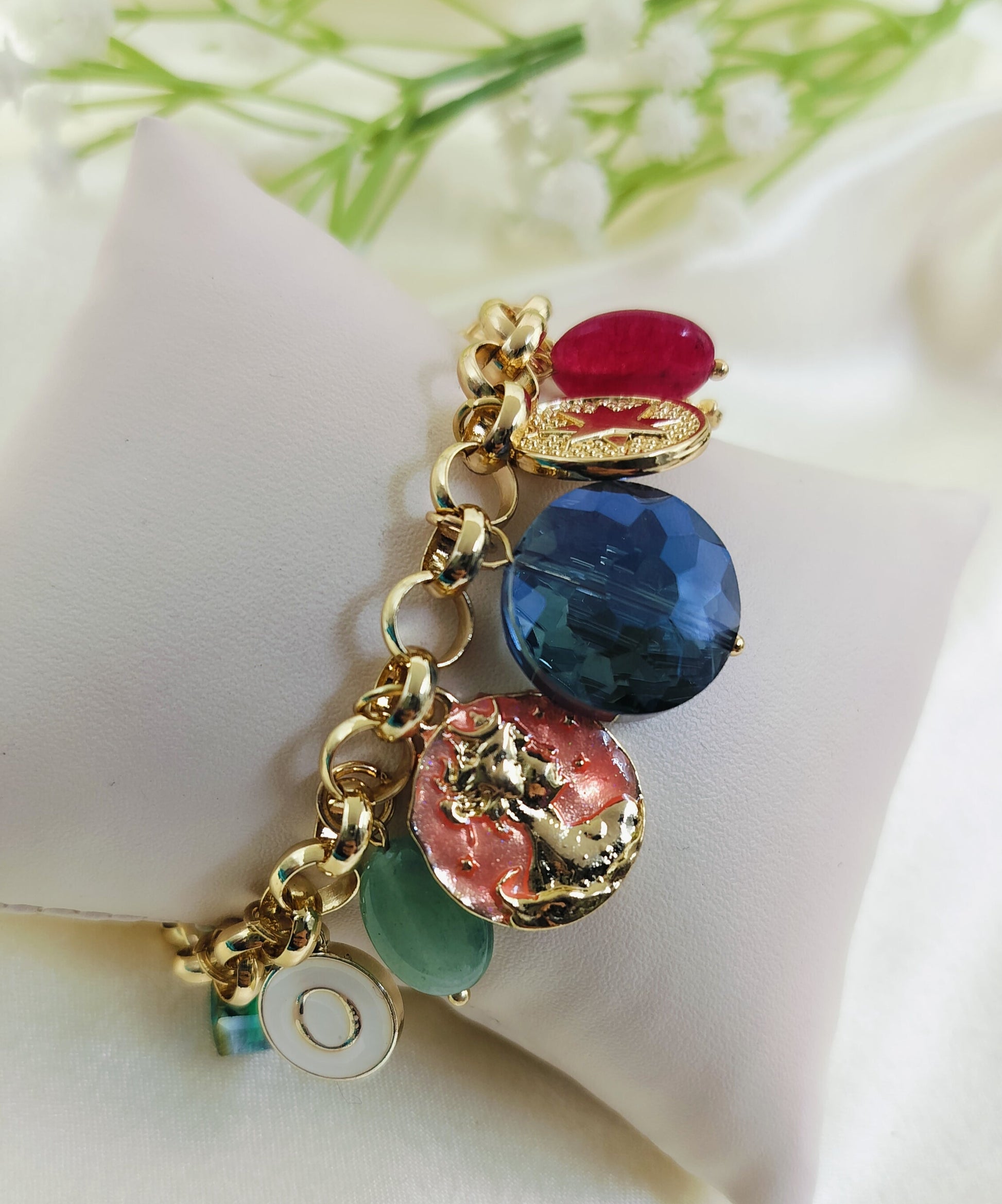Gold chain bracelet with multicolor gemstone charms in red, blue, green, and pink, featuring an embossed coin-style pendant, stylish women’s jewelry on fabric cushion background