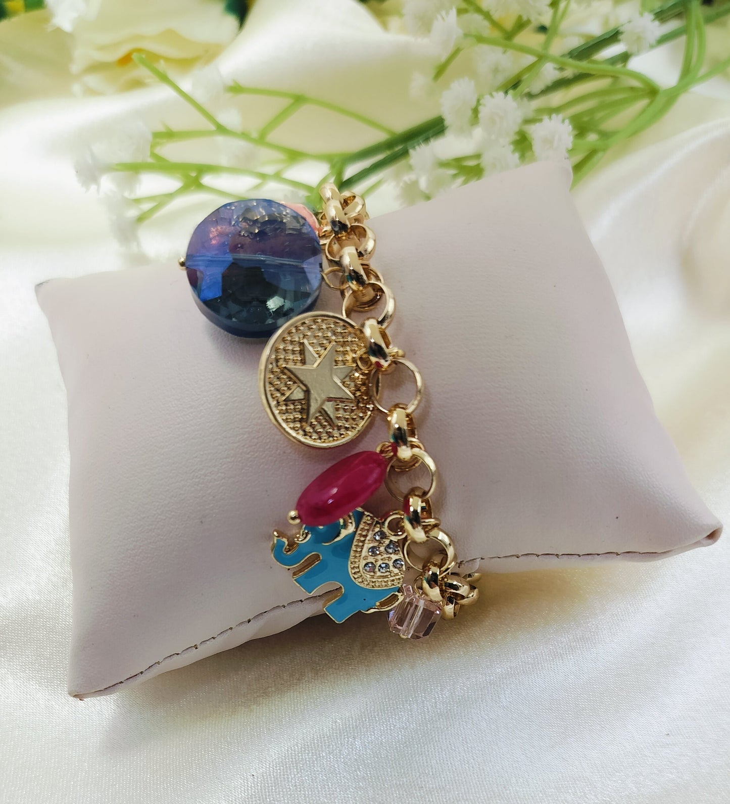 Gold chain bracelet with bold blue gemstone charm, embossed star coin pendant, red bead, and enamel elephant accent, fashionable women’s jewelry on satin fabric cushion background