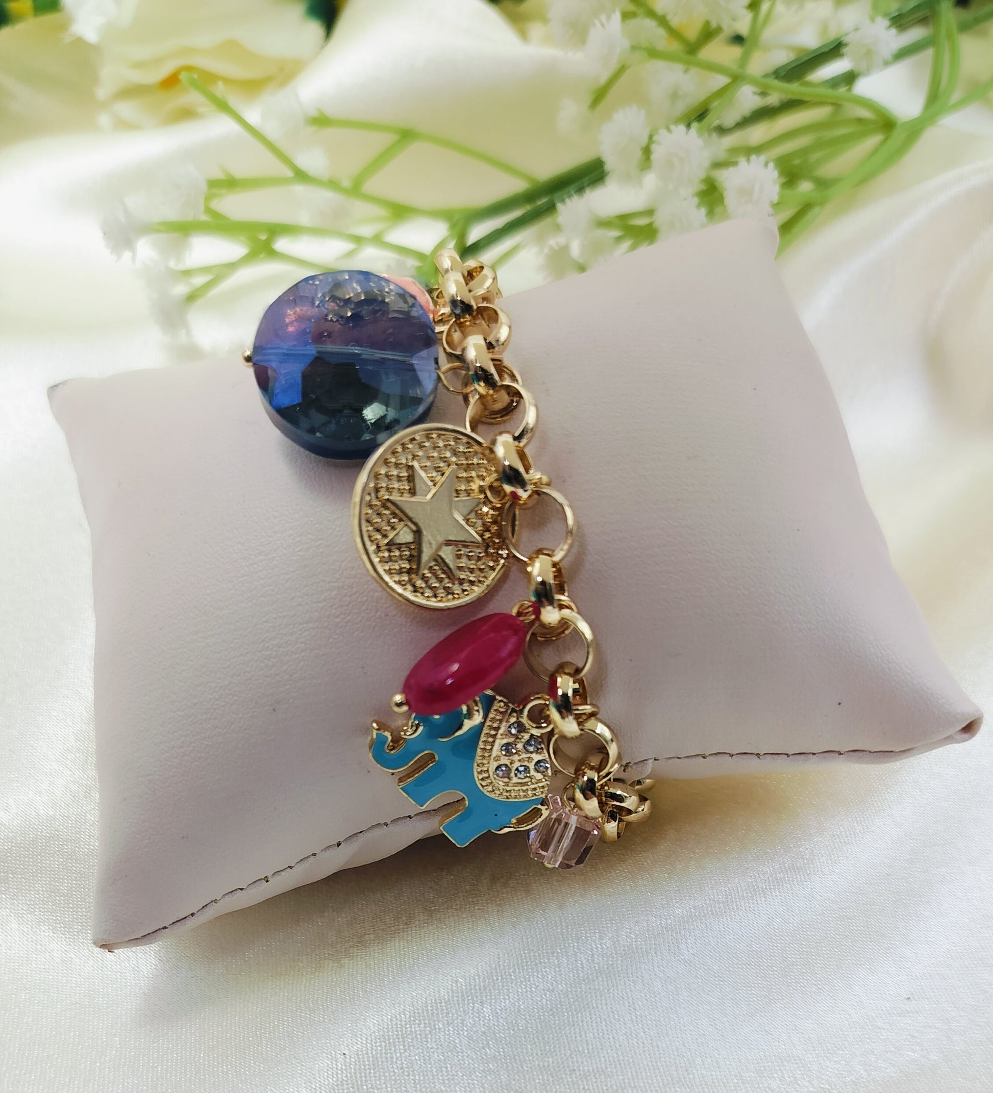 Gold chain bracelet with bold blue gemstone charm, embossed star coin pendant, red bead, and enamel elephant accent, fashionable women’s jewelry on satin fabric cushion background
