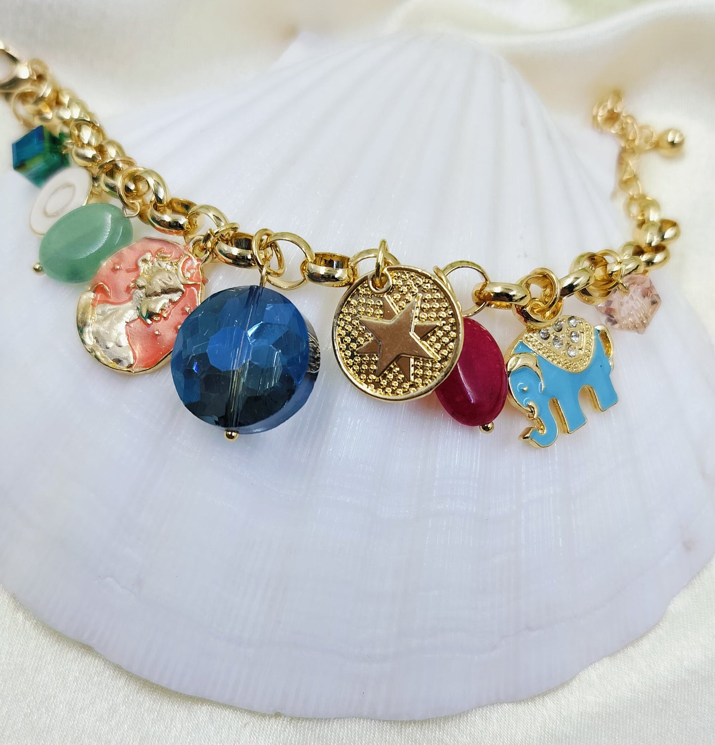 A gold-colored charm bracelet with a variety of colorful charms, displayed on a white seashell.