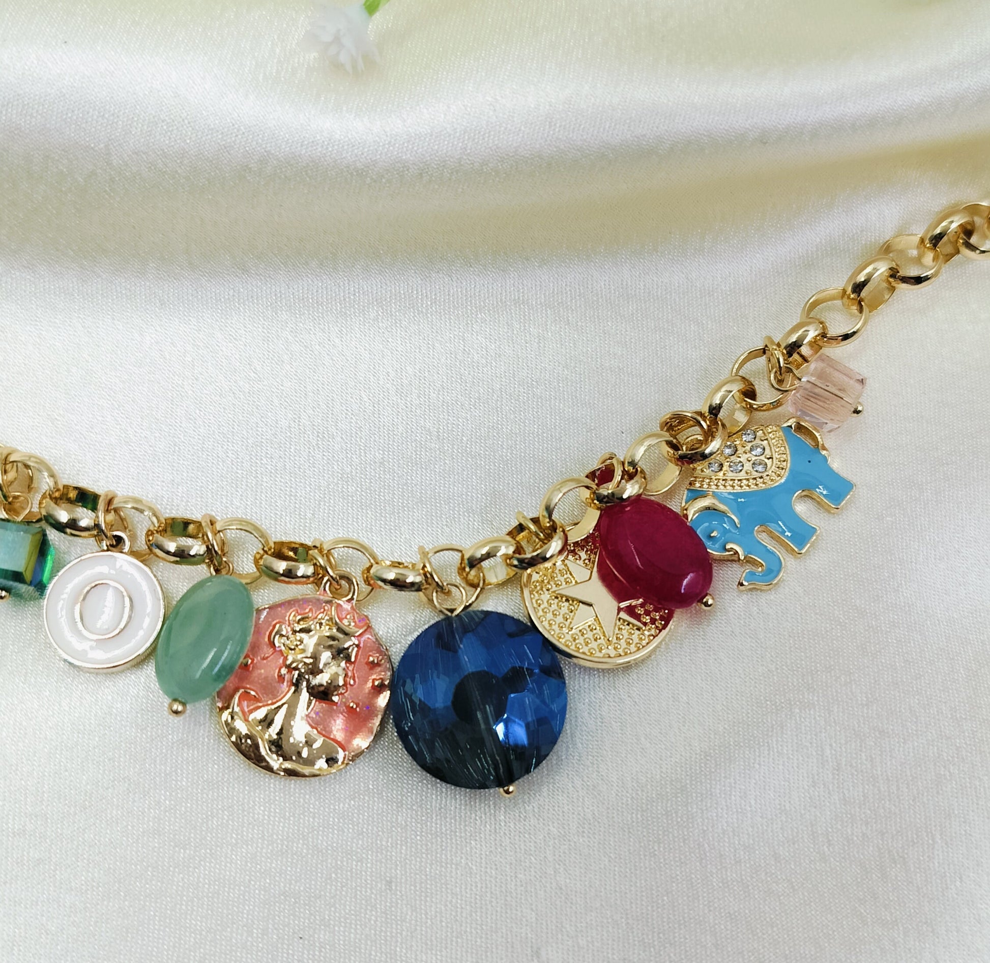 Gold chain bracelet with colorful charms including blue gemstone, red stone, green bead, embossed coin pendant, enamel elephant, and round accents, trendy women’s jewelry on satin fabric background
