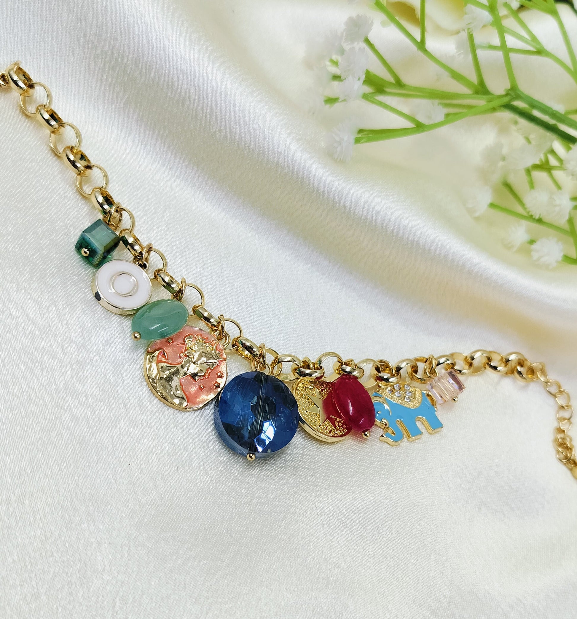 Gold chain bracelet with assorted charms including green bead, white round accent, embossed coin pendant, blue gemstone, red stone, and enamel elephant, stylish women’s jewelry on satin fabric background
