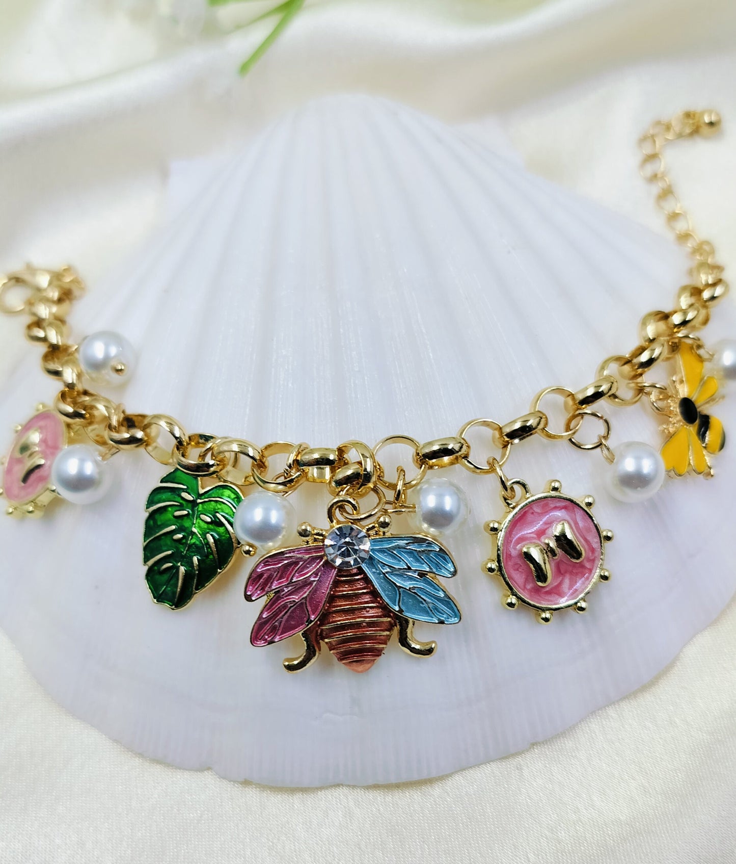 A gold bracelet with pearl accents and various charms including a bee, butterfly, and monstera leaf, displayed on a white surface.