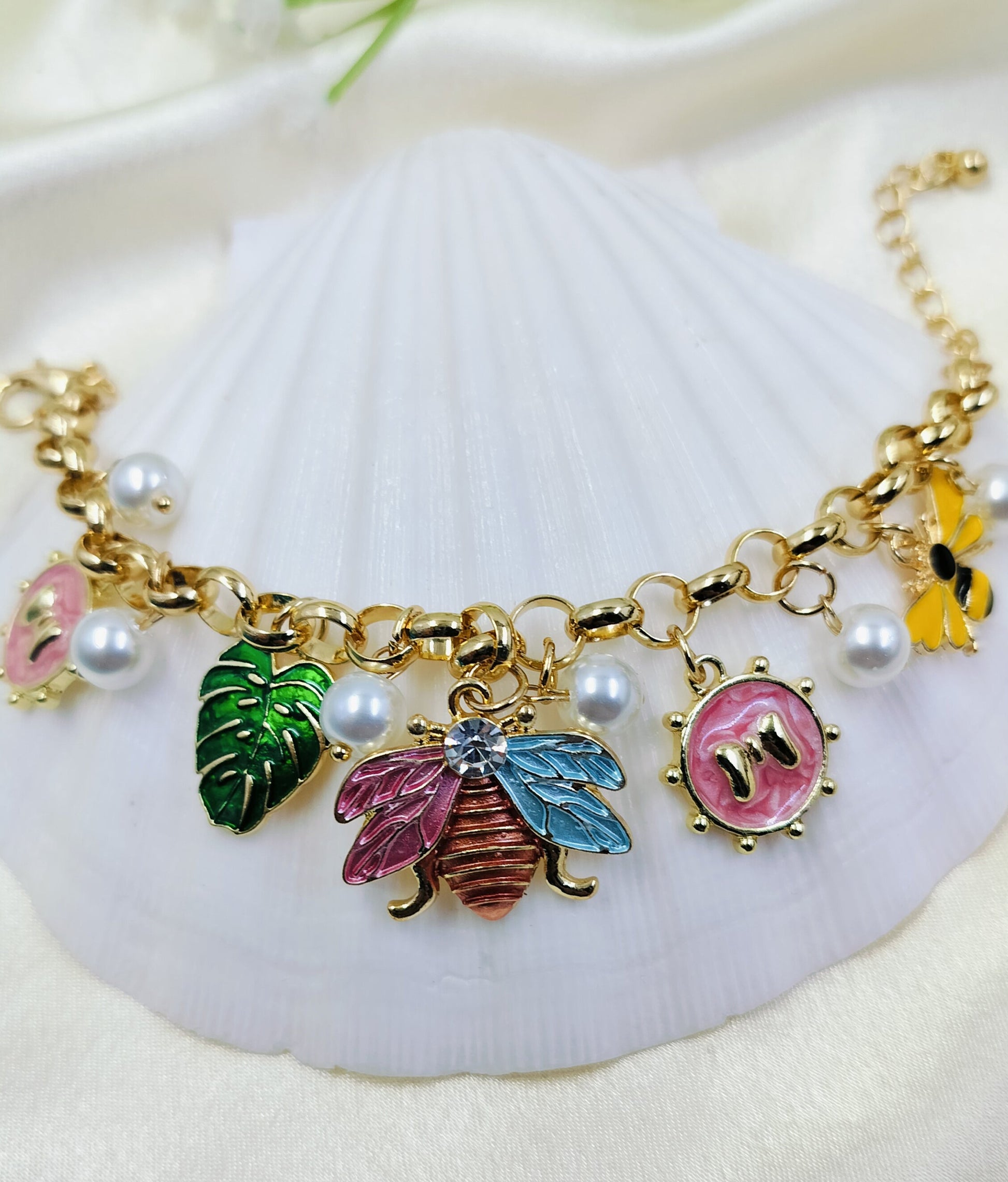 A gold bracelet with pearl accents and various charms including a bee, butterfly, and monstera leaf, displayed on a white surface.