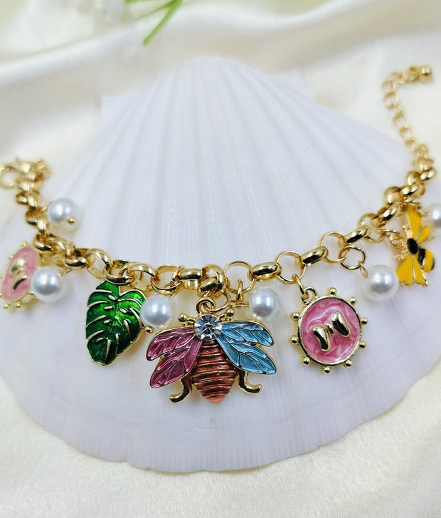 A gold bracelet with pearl accents and various charms including a bee, butterfly, and monstera leaf, displayed on a white surface.