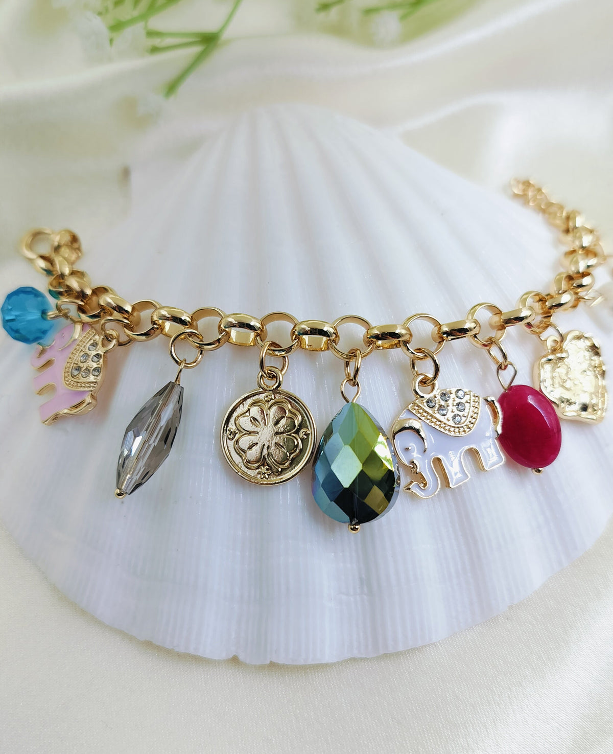 A gold charm bracelet featuring embellished elephant charms, a four-leaf clover medallion, and faceted gemstone drops, displayed on a white surface.
