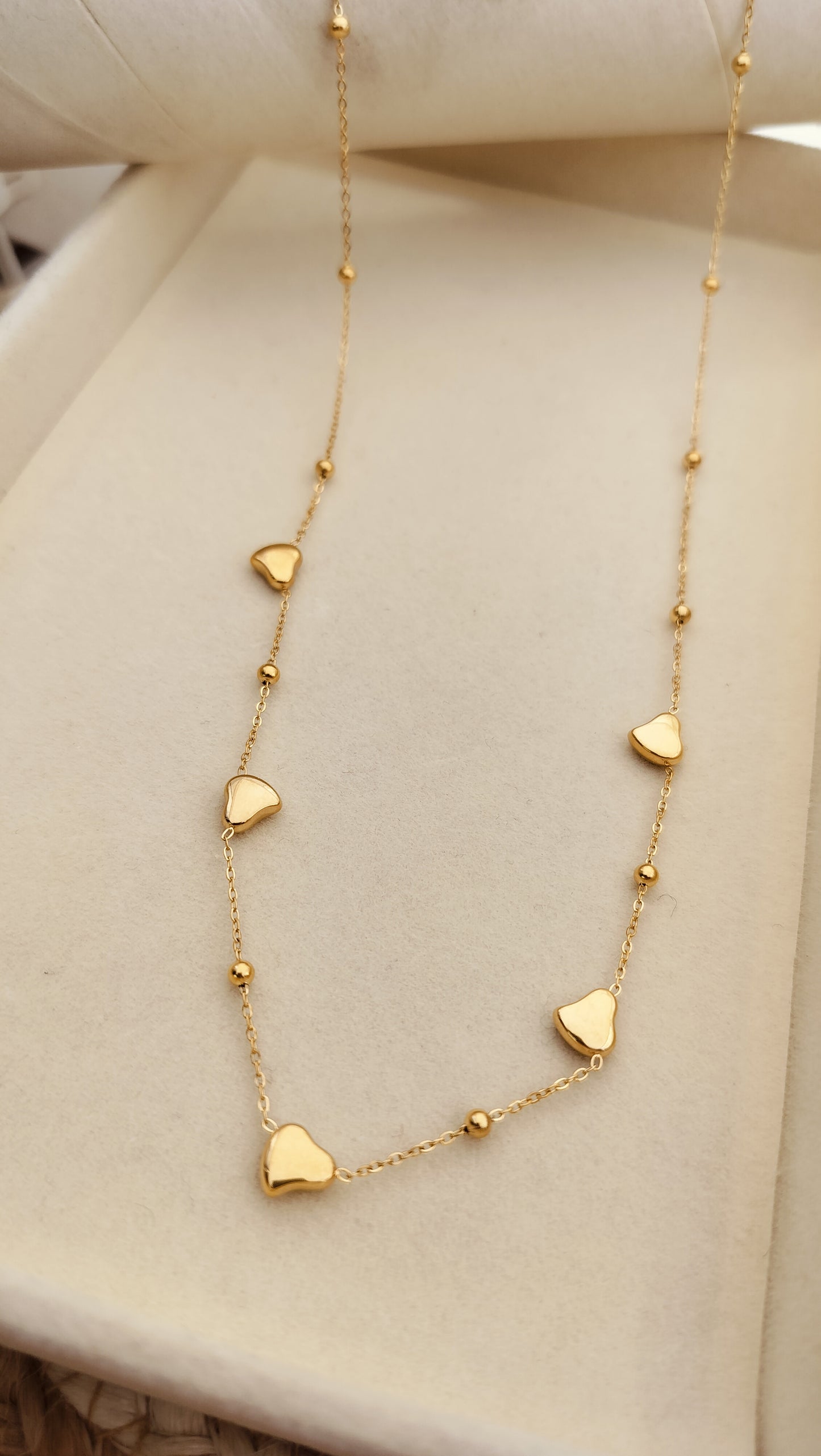 A gold necklace with linked heart charms and subtle gold bead accents displayed on a beige background.