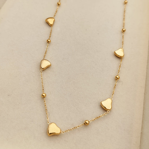 A gold necklace with linked heart charms and subtle gold bead accents displayed on a beige background.
