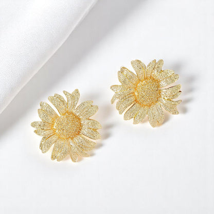 Golden sunflower-shaped earrings displayed on a plain white background with a folded cloth beside them.