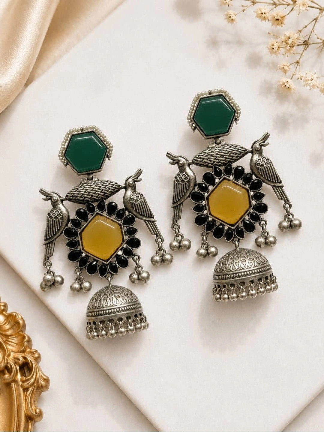 Pair of oxidised silver bird jhumka earrings with green hexagonal studs and yellow centre stones laid flat on white surface