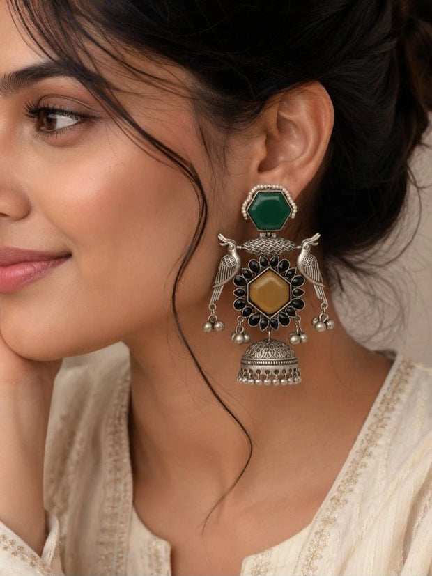 Woman wearing large oxidised silver bird jhumka earring with green stud top and yellow stone against a cream kurta