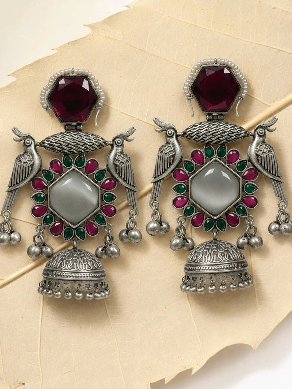 Pair of oxidised silver peacock jhumka earrings with red and green stones laid flat on a textured cream surface