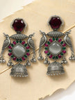 Pair of oxidised silver peacock jhumka earrings with red and green stones laid flat on a textured cream surface