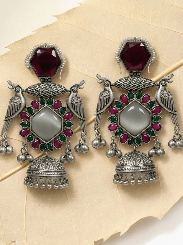 Pair of oxidised silver peacock jhumka earrings with red and green stones laid flat on a textured cream surface